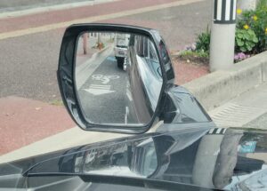 車のフェンダーミラーに映る自転車レーンと後続車の様子。路肩の赤い歩道や縁石、電柱が写り、巻き込み確認の重要性を示す実写写真。