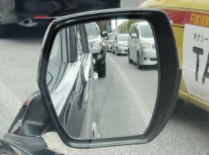 車のフェンダーミラーに映る渋滞中の後続車列。手前には福岡のタクシー車両が写り、交通状況を確認するリアルな運転視点の写真。