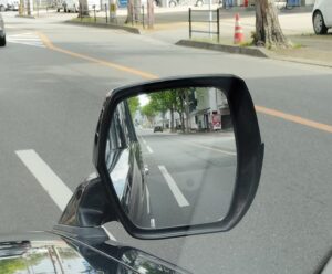 車のフェンダーミラーに映る街中の直進道路。白い車線、街路樹、店舗が並び、前方と後方の状況を同時に把握する運転視点の実写写真。